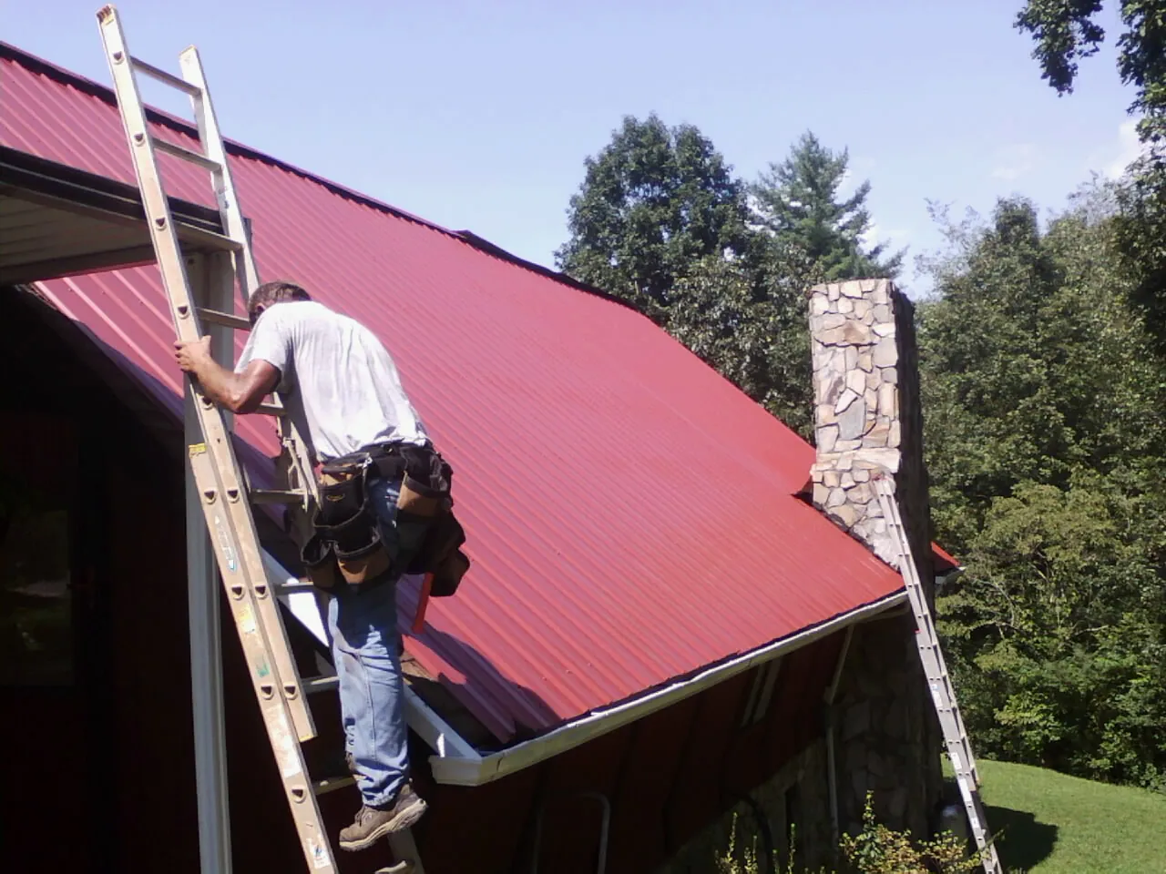Roofer on a ladder beside a red metal roof and stone chimney — Soffit & Fascia Repair by Roofing Brothers in Red Lion